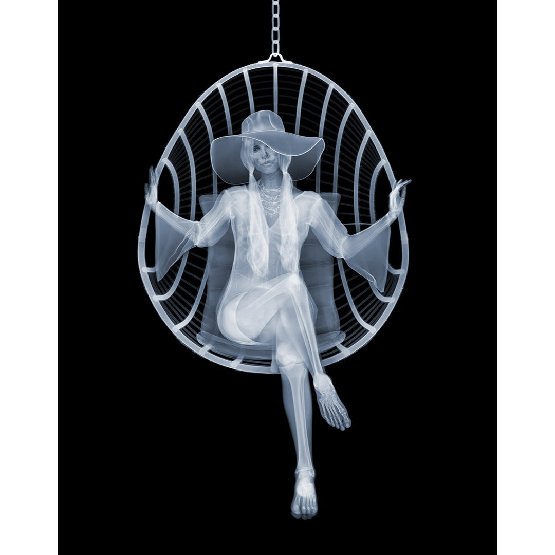 Framed Chromaluxe black and white x-ray photograph by Nick Veasey of a skeleton woman sitting in a swing chair with long pigtails, large hat and boho style dress crossing her legs