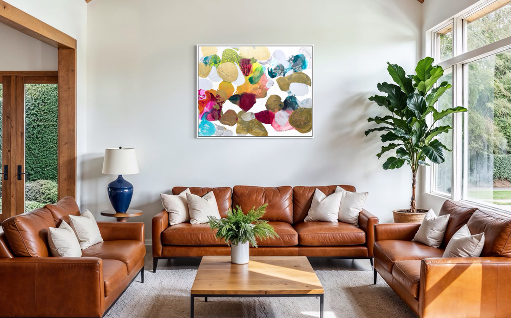 Liz Barber, Butterflies 3, acrylic paint, ink, and watercolor on canvas, 60 x 48 inches. Abstract composition of rounded shapes in metallic gold, olive, crimson, magenta, orange, yellow, turquoise, teal, and white on a white background, shown in a white frame in a living room with cognac leather sofas, a wood coffee table, and a fiddle-leaf fig by large windows.