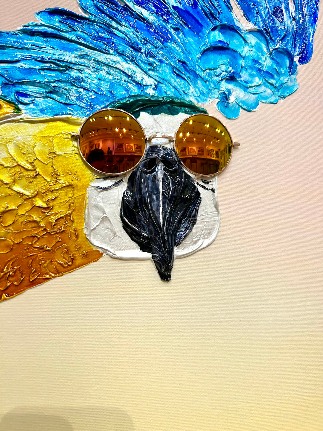 30” x 30” painting by French artist Moogly of a blue and yellow parrot in sunglasses with a pink and yellow ombre background. The parrot is depicted in mid-flight and made out of resin, giving it a textured and shiny finish. The sunglasses are real round, reflective sunglasses that the artist has adhered to the canvas.