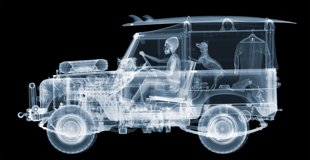 Framed Chromaluxe black and white x-ray photograph by Nick Veasey of a skeleton man driving a land rover with a surf board on the roof and a dog and surf suit in the back of the car. 