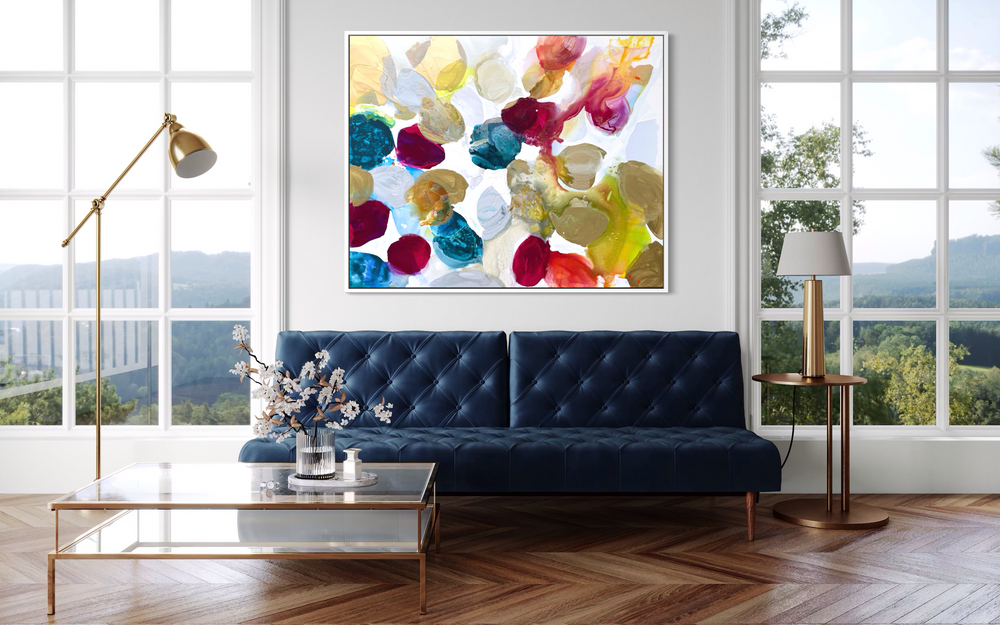 Liz Barber, Spring Dream, acrylic paint, ink, and watercolor on canvas, 60 x 48 inches. Abstract composition of rounded shapes in gold, taupe, deep crimson, teal, pale blue, and white with red-orange and chartreuse ink washes on a white background, shown in a white frame above a navy tufted sofa with a brass floor lamp and glass coffee table in front of large windows.
