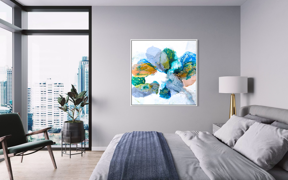 Liz Barber, Water's Edge 1, acrylic paint, ink, and watercolor on canvas, 48 x 48 inches. Abstract composition of overlapping rounded, stone-like shapes in teal, cobalt blue, lavender gray, ochre, and peach with speckled yellow-green texture on a white background, shown in a white frame above a bed in a gray bedroom with floor-to-ceiling windows overlooking city high-rises.